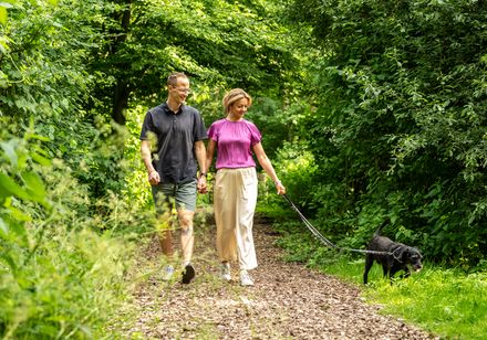 Ein Paar geht mit einem schwarzen Hund auf einem Naturweg durch dichte grüne Vegetation spazieren.