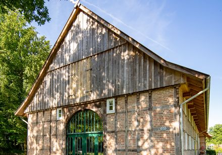 Altes Backsteingebäude mit Fachwerkelementen, umgeben von grüner Wiese und Bäumen im Sonnenschein.