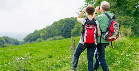 Ein Paar mit Wanderrucksäcken blickt von einer grünen Wiese aus mit Ferngläsern in die Ferne.