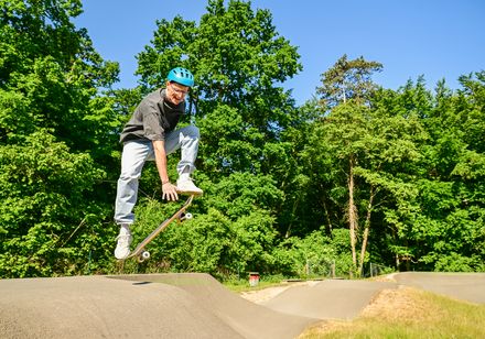 Ein Skateboarder mit blauem Helm springt auf einem Pumptrack vor üppigem grünen Wald im Sonnenlicht.
