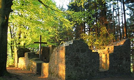 Ruinen der Tönsbergkapelle im Wald mit Kreuz, von Sonnenlicht beleuchtet, umgeben von Bäumen.