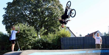 BMX-Fahrer dreht einen Rückwärtssalto über dem Pumptrack, zwei Zuschauer filmen am Rand.