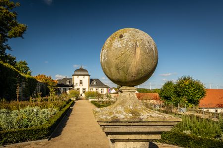 Eine historische Sonnenuhr aus Stein in einem gepflegten Schlossgarten vor einem barocken Gebäude.