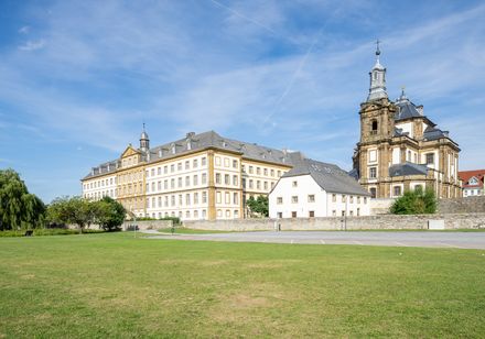 Barockes Kloster mit mächtigem Turm und weitläufiger Rasenfläche unter blauem Himmel.