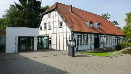 Historisches Fachwerkgebäude mit modernem Anbau, umgeben von gepflastertem Vorplatz und grünem Garten.