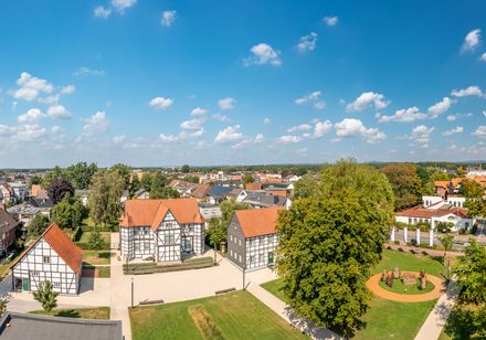 Dorfpanorama mit Fachwerkhäusern, Bäumen und modernen Gebäuden unter blauem Himmel mit Wolken.