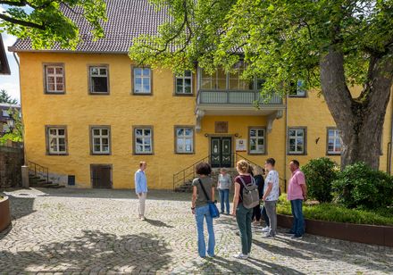 Ein historische Gebäude mit gelber Fassade und kleinen Sprossenfenstern. Menschen stehen davor.