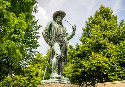 Bronzestatue eines Mannes in traditioneller Tracht mit Pfeife, umgeben von grünen Bäumen.