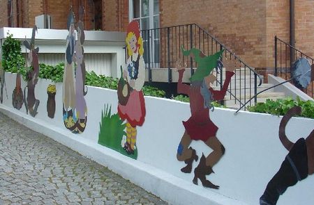 Bunte Märchenfiguren schmücken die weiße Mauer vor einem Backsteingebäude mit großen Fenstern.