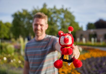 Ein Mann hält freudig ein rotes Stofftier in Form eines Käfers, umgeben von bunten Blumen.