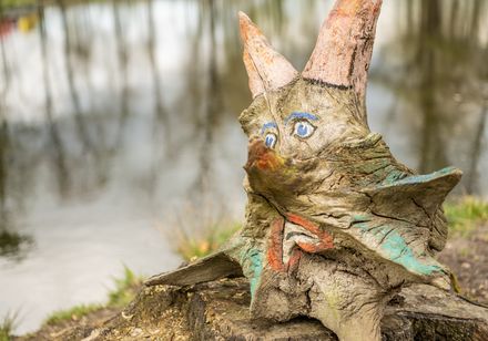 Eine skurrile, bemalte Baumwurzel mit Gesichtsmerkmalen und Hörnern am Ufer eines ruhigen Sees.