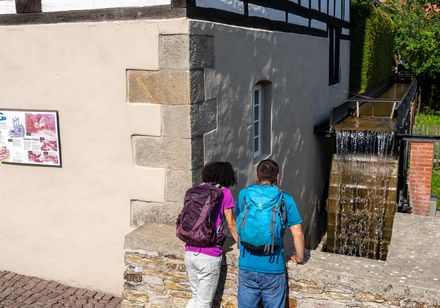 Zwei Personen mit Rucksäcken betrachten eine alte Mühle mit fließenden Wasserkaskaden.