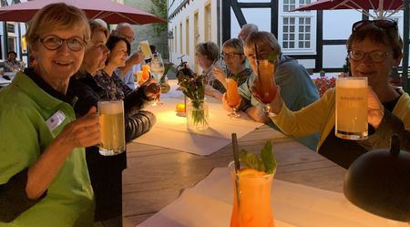 Gruppe älterer Menschen sitzt unter Sonnenschirmen an langen Tischen und genießt bunte Getränke.