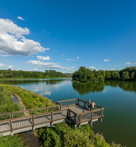 <p>Eine idyllische Flusslandschaft mit Aussichtsplattform und zwei Menschen, umgeben von üppiger Natur.</p>