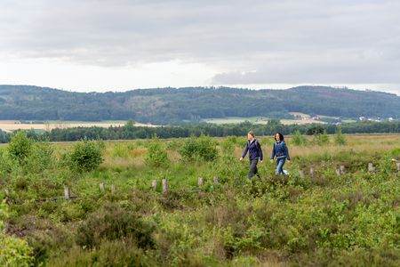 Zwei Wanderinnen spazieren durch eine weitläufige, grüne Heidelandschaft unter bewölktem Himmel.