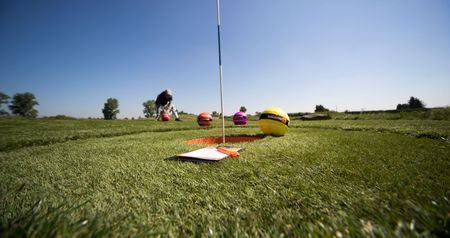 Mehrere bunte Fußbälle auf einem Golfplatzrasen nahe eines Lochs mit Flagge, klarer Himmel im Hintergrund.