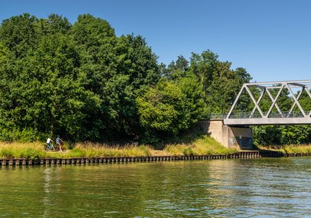 Radfahrer neben grünem Kanal, Eisenbahnbrücke darüber, umgeben von üppigen Bäumen, blauer Himmel.