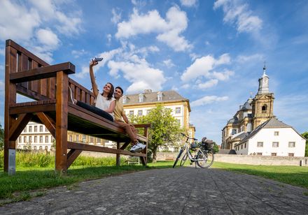 Paar auf großer Holzbank macht Selfie vor historischem Gebäude mit Kirche, Fahrräder im Vordergrund.