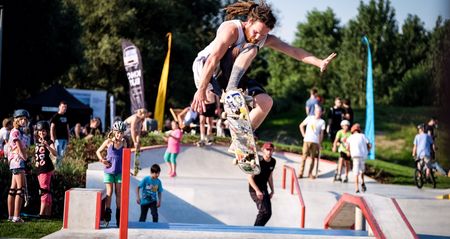 Ein Skateboarder führt in einem lebhaften Skatepark bei sonnigem Wetter einen Sprungtrick aus.