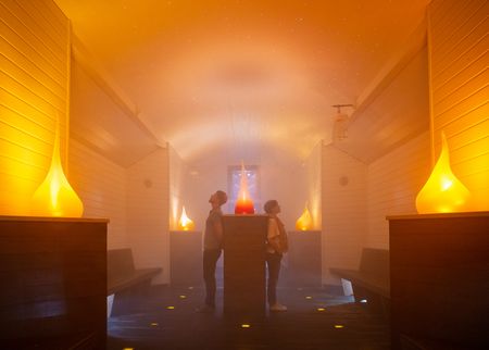 Zwei Personen stehen in einem stimmungsvoll beleuchteten, mit Nebel gefüllten Raum mit hölzernem Interieur.