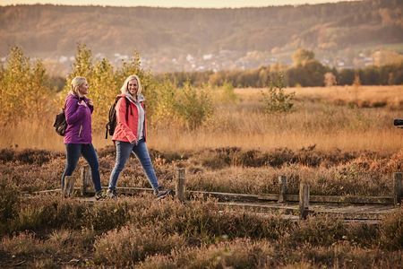 Zwei Frauen wandern im Herbst bei Sonnenuntergang durch eine malerische Heidelandschaft.