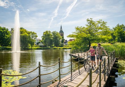 Ein Paar mit Fahrrädern spaziert auf einem Steg über einen See, umgeben von Natur und einem Kirchturm.