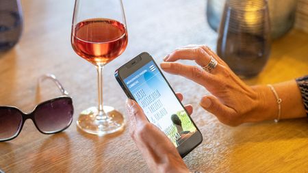 Eine Hand bedient ein Smartphone neben einem Glas Wein auf einem Tisch, mit einer Sonnenbrille daneben.