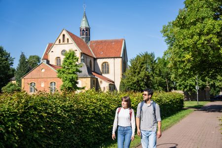 Zwei Personen spazieren auf einem Weg entlang einer begrünten Hecke, im Hintergrund eine historische Kirche.