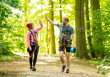 Zwei Personen im Kletterpark geben sich ein High-Five, umgeben von grünem Wald.