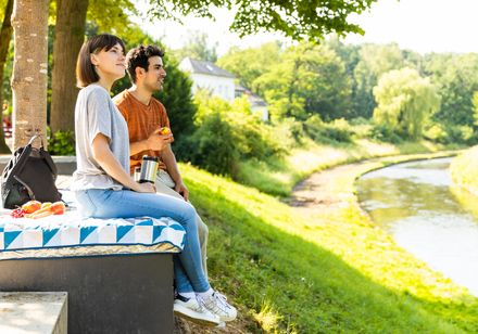 Ein Paar sitzt auf einer Bank am Flussufer, genießt den sonnigen Tag und picknickt entspannt.