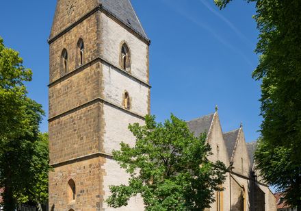 Eine alte Kirche aus hellem Stein mit hohem Turm, umrahmt von grünem Laub und blauem Himmel.