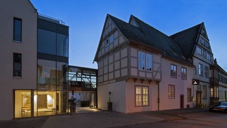 Abendliche Außenansicht eines modernen Gebäudetrakts neben einem historischen Fachwerkhaus.