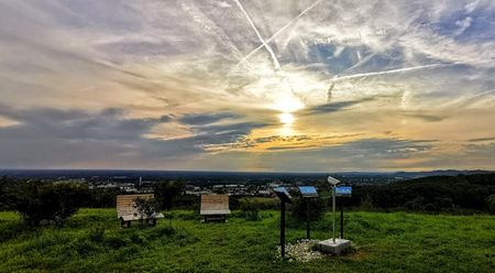 Sonnenuntergang über einer grünen Landschaft mit Info-Tafeln und Parkbänken unter dramatischem Himmel.