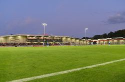 Fußballstadion in Abendstimmung, Flutlicht erleuchtet das Spielfeld, Zuschauer auf Tribünen.