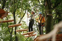 Zwei Personen mit Helmen und Kletterausrüstung überqueren eine schwebende Holzbrücke im Kletterpark.