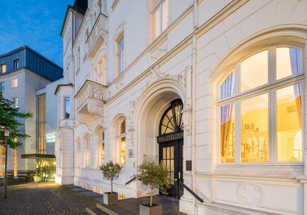 Außenansicht eines stilvollen, historischen Hotelgebäudes mit heller Fassade, großen Fenstern und beleuchtetem Eingangsbereich in zentraler Lage.