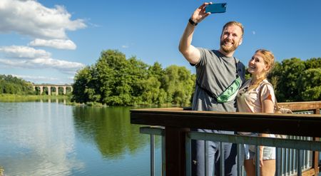 Ein lächelndes Paar macht auf einer Brücke ein Selfie mit einem Fluss und Bäumen im Hintergrund.