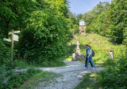 Ein Paar spaziert auf einem Waldweg vorbei an einem neoklassischen Denkmal im sommerlichen Grün.