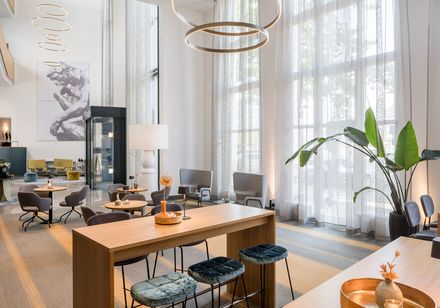 Großzügiger, modern gestalteter Loungebereich mit hohen Fenstern, stilvollen Sitzgruppen und dekorativer Beleuchtung.
