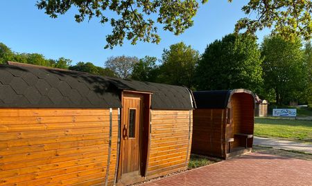 Zwei hölzerne Campingfässer stehen nebeneinander auf einem gepflasterten Weg, umgeben von Bäumen.