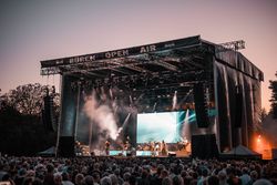 Großes Freiluftkonzert bei Sonnenuntergang mit Künstlern auf der Bühne und Publikum im Vordergrund.