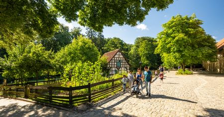 Eine Familie spaziert über Kopfsteinpflaster an einem Fachwerkhaus entlang, umgeben von grünen Bäumen.