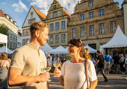 Menschen genießen Wein beim Sommerfest in historischer Altstadt mit Zelten und sonnigem Wetter.