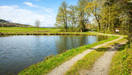 Teich, rechts ein Weg und im Hintergrund eine Infotafel
