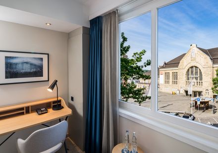 Helles Hotelzimmer mit Schreibtisch und großem Fenster, das einen Blick auf ein historisches Bahnhofsgebäude und den Vorplatz bietet.