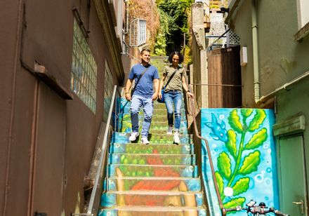 Zwei Menschen gehen eine bunt bemalte Treppe hinunter, entlang einer schmalen Gasse bei sonnigem Wetter.