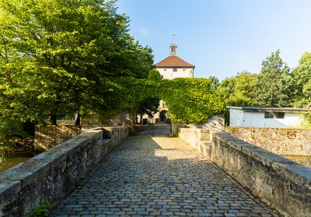 Zugang über eine historische, mit Kopfsteinpflaster belegte Brücke zu einem Schlosstor im Grünen.