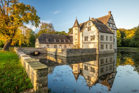 Wasserschloss umgeben von herbstlichen Bäumen, spiegelnd im ruhigen Gewässer des Grabens.