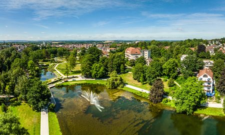 Der Bild zeigt einen weitläufigen Park mit grünen Wiesen, einem Teich und einem zentralen Wasserbrunnen.