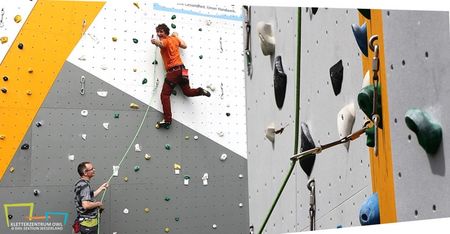 Kletterer erklimmt bunte Kletterwand, abgesichert durch eine Person am Boden, die Seil sichert.